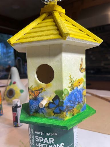 Birdhouse decorated with flowers and butterflies, featuring a yellow roof and green base, sitting on a can of varnish.