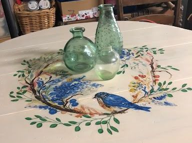 Round table with painted bluebird and greenery, three green glass bottles.