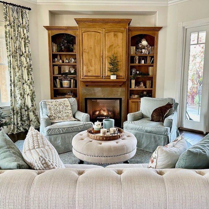 Cozy living room with a fireplace, built-in shelves, and light upholstered chairs around a round ottoman.