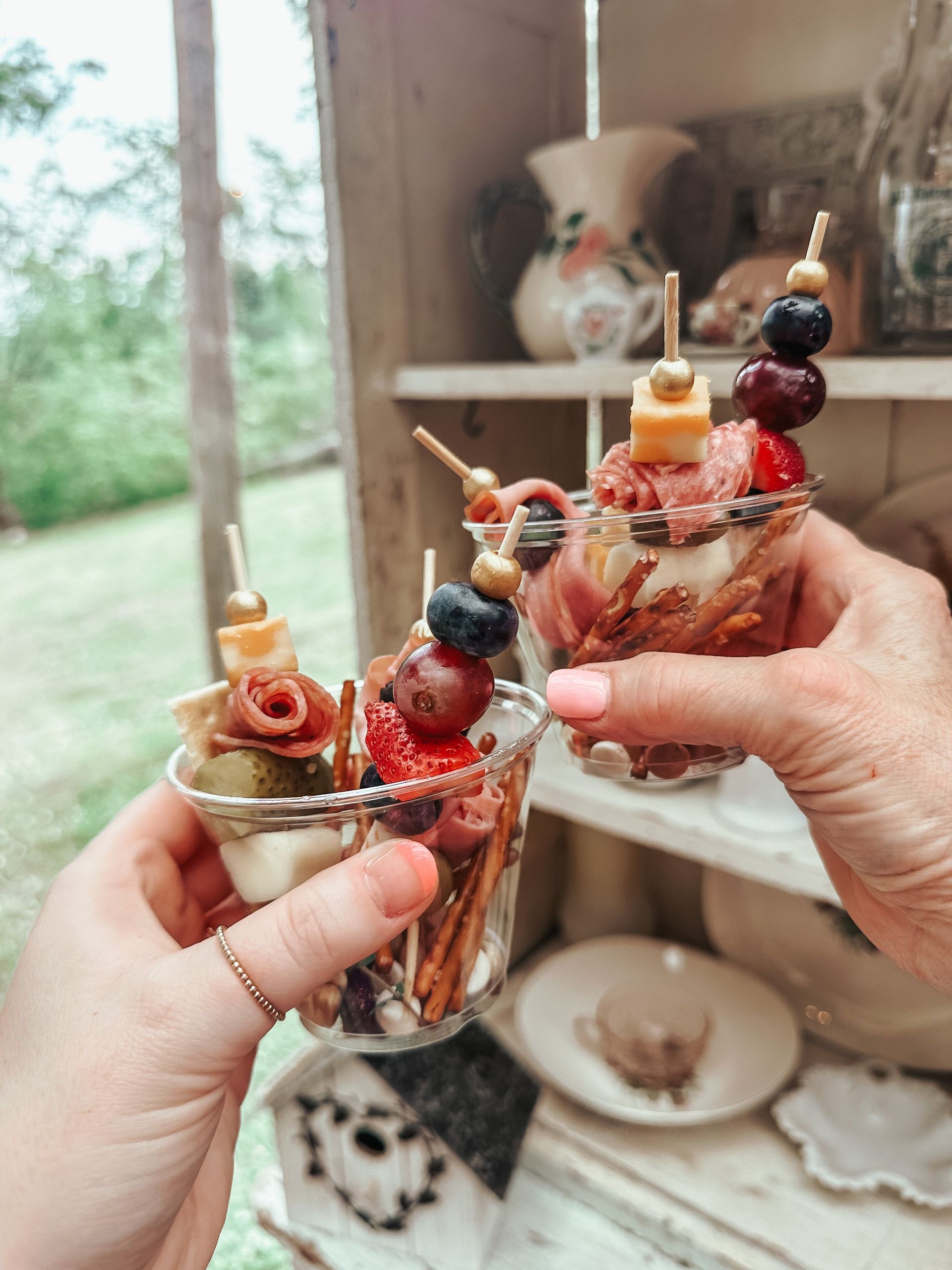 Two hands holding charcuterie cups with skewers, outdoors.