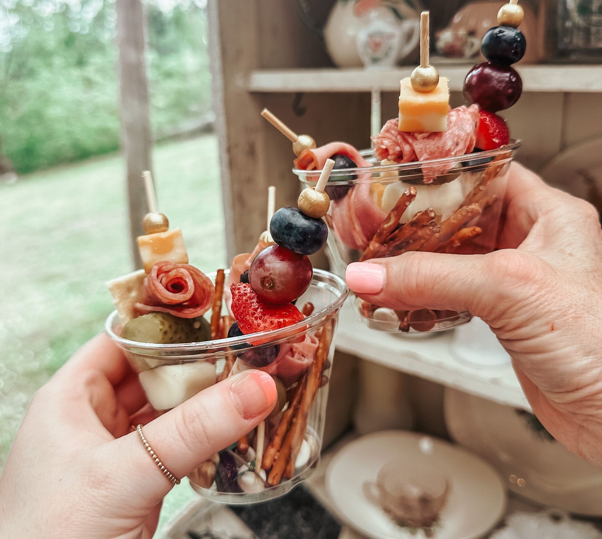 Two hands holding charcuterie cups with skewers, assorted meats, cheese, and fruit.