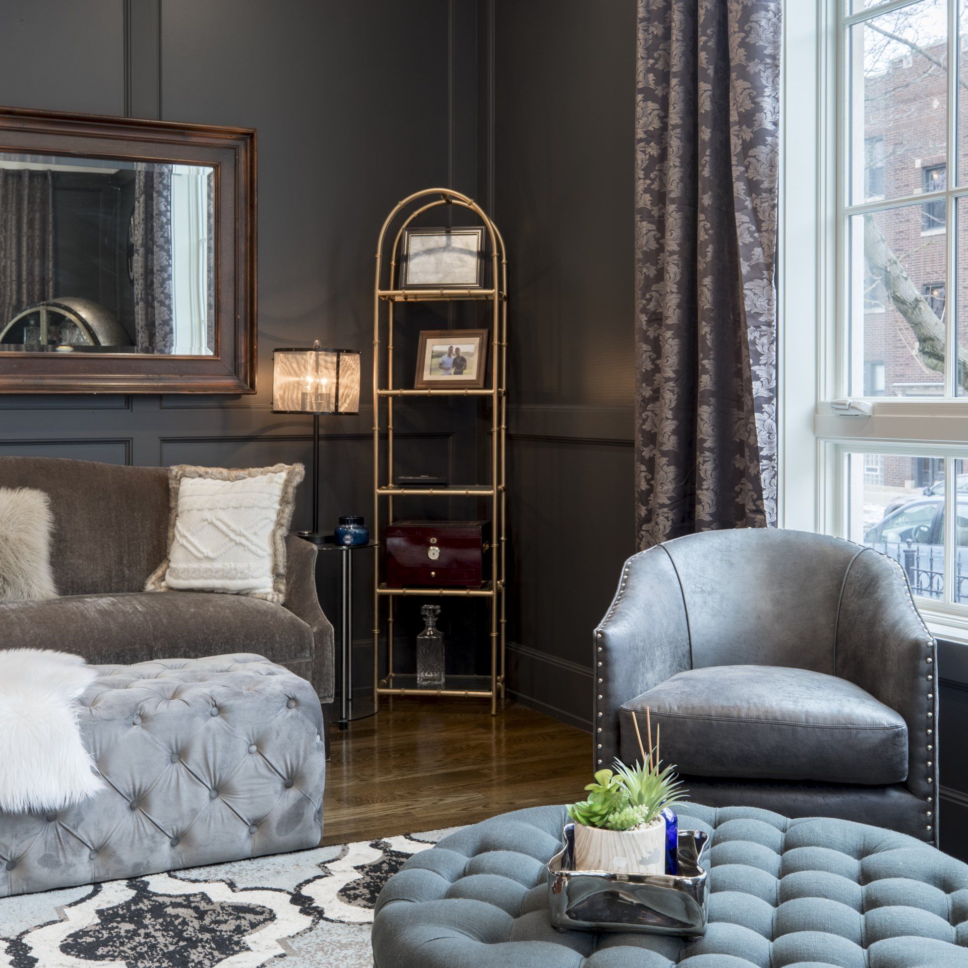 Cozy living room with dark gray walls, sofa, armchair, gold shelving unit, and round ottoman.