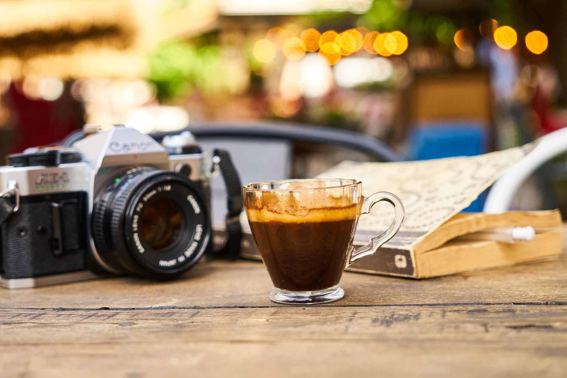 Espresso in glass cup, vintage camera, and map on a wooden table, blurred outdoor background.