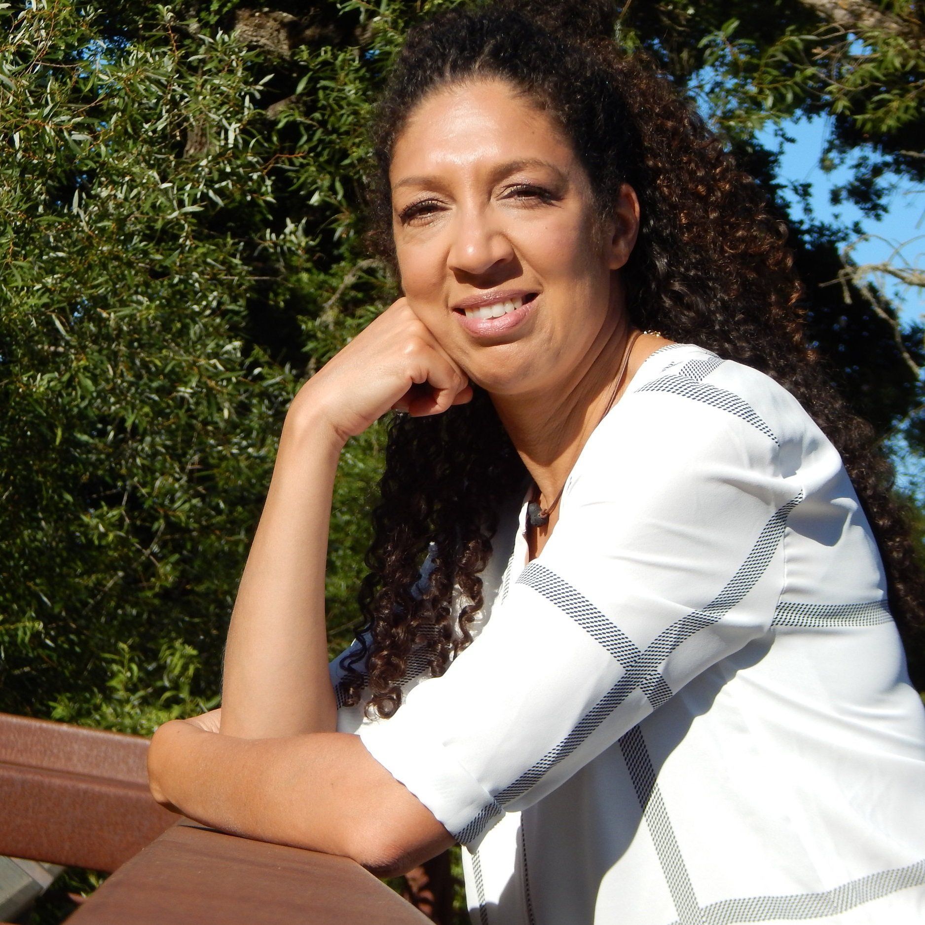 Woman with long curly hair smiles, resting arm on a wooden rail outdoors. Wearing white top.