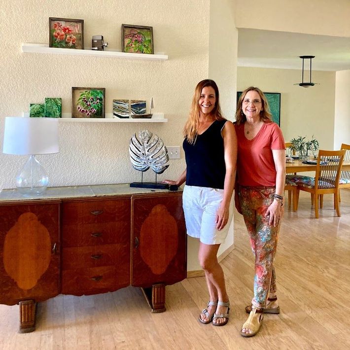 Two women standing in a bright living room beside a wooden sideboard with shelves and decor.