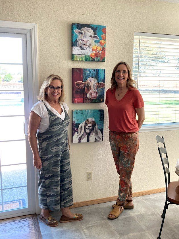Two women stand by three colorful cow paintings on a wall.