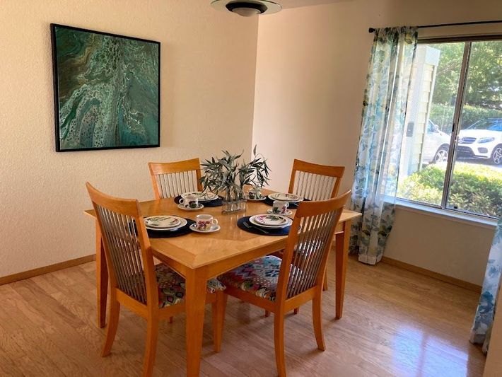 Dining room with a wooden table set for four beside a large window and a wall painting.