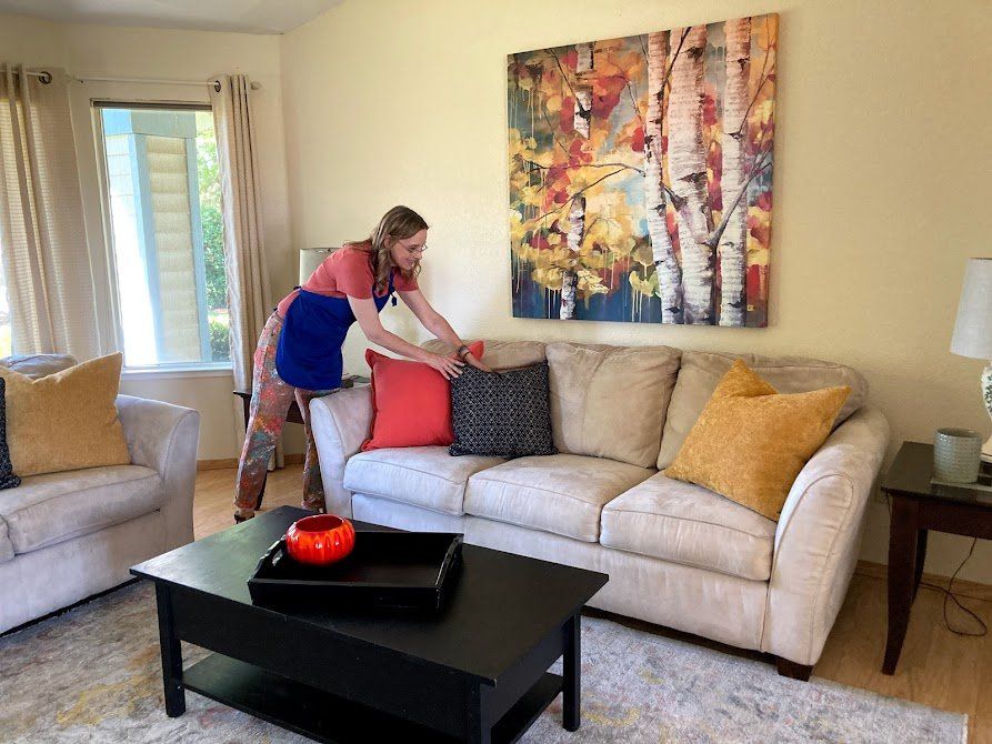 Woman arranging pillows on a beige sofa in a living room, with a coffee table and art on the wall.