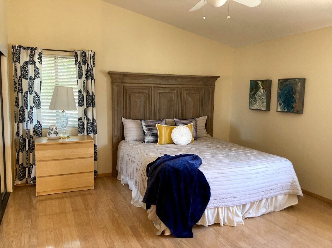 Bedroom with wooden floor, bed with light bedding, wooden headboard, yellow pillows, blue throw, and decorative curtains.