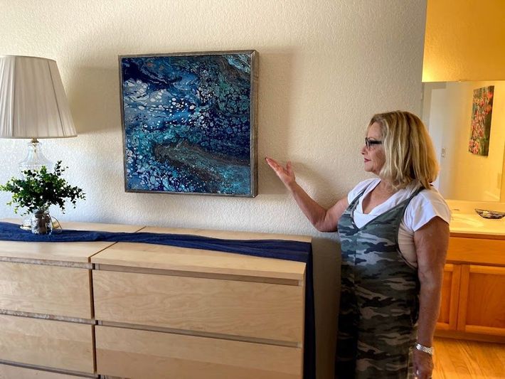 Woman in a home living room points at blue abstract wall art above a wooden dresser.
