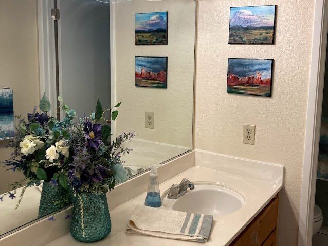 Bathroom vanity with artwork and floral arrangement. White countertop, round sink, and small paintings on wall.