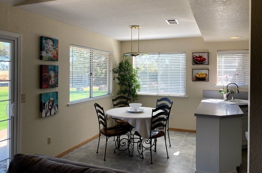 Dining area with round table, four chairs, artwork, windows with blinds.