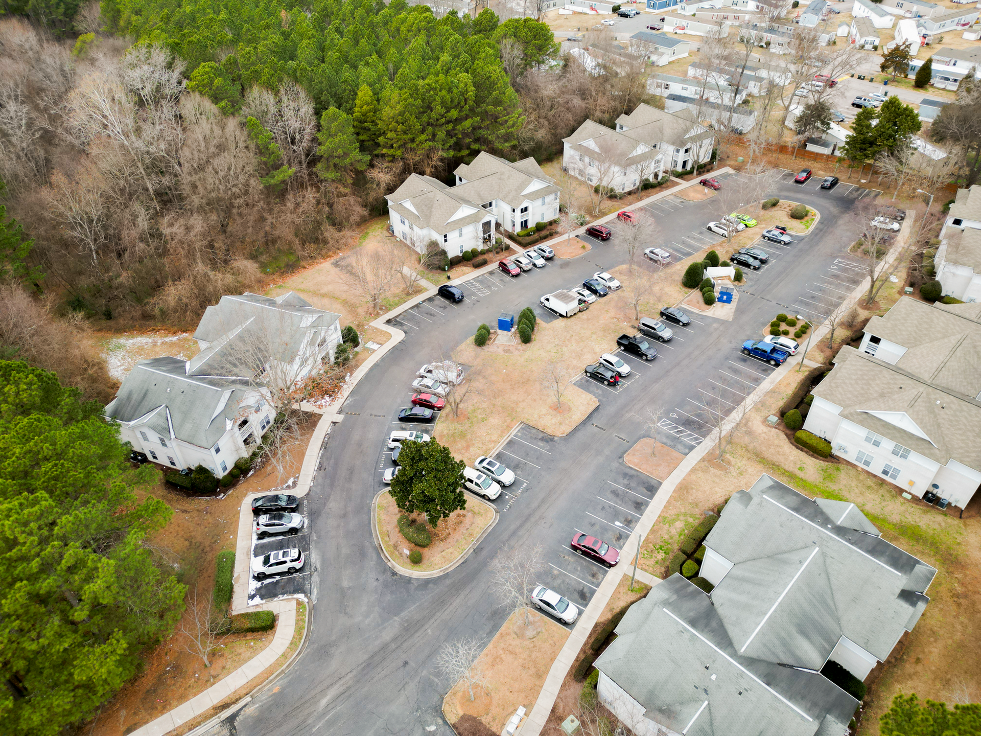 Aerial view of apartments with parking lot and cars; surrounded by trees and residential area.