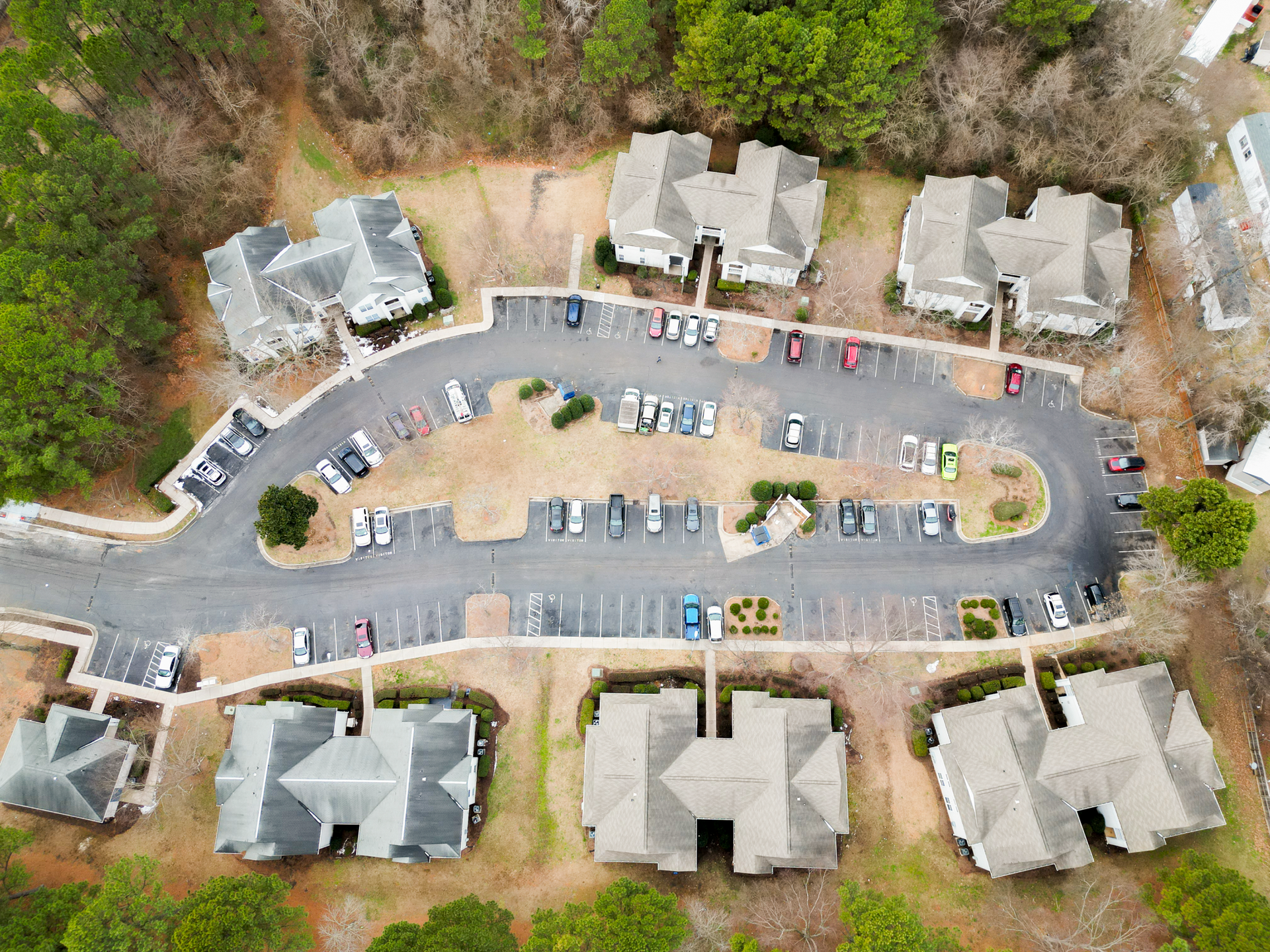 Aerial view of apartment complex with parking lot, cars, and surrounding trees. Gray roofs, tan asphalt.