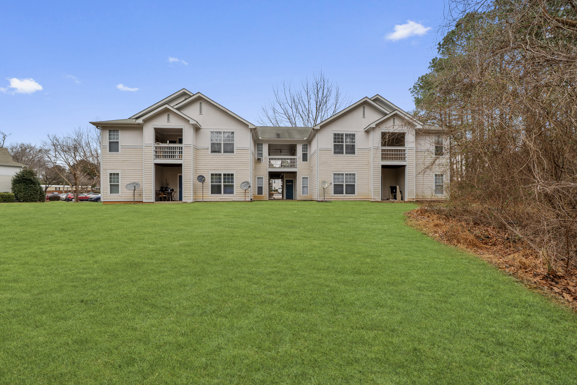 Two-story apartment building with beige facade, balconies, and a large green lawn under a blue sky.