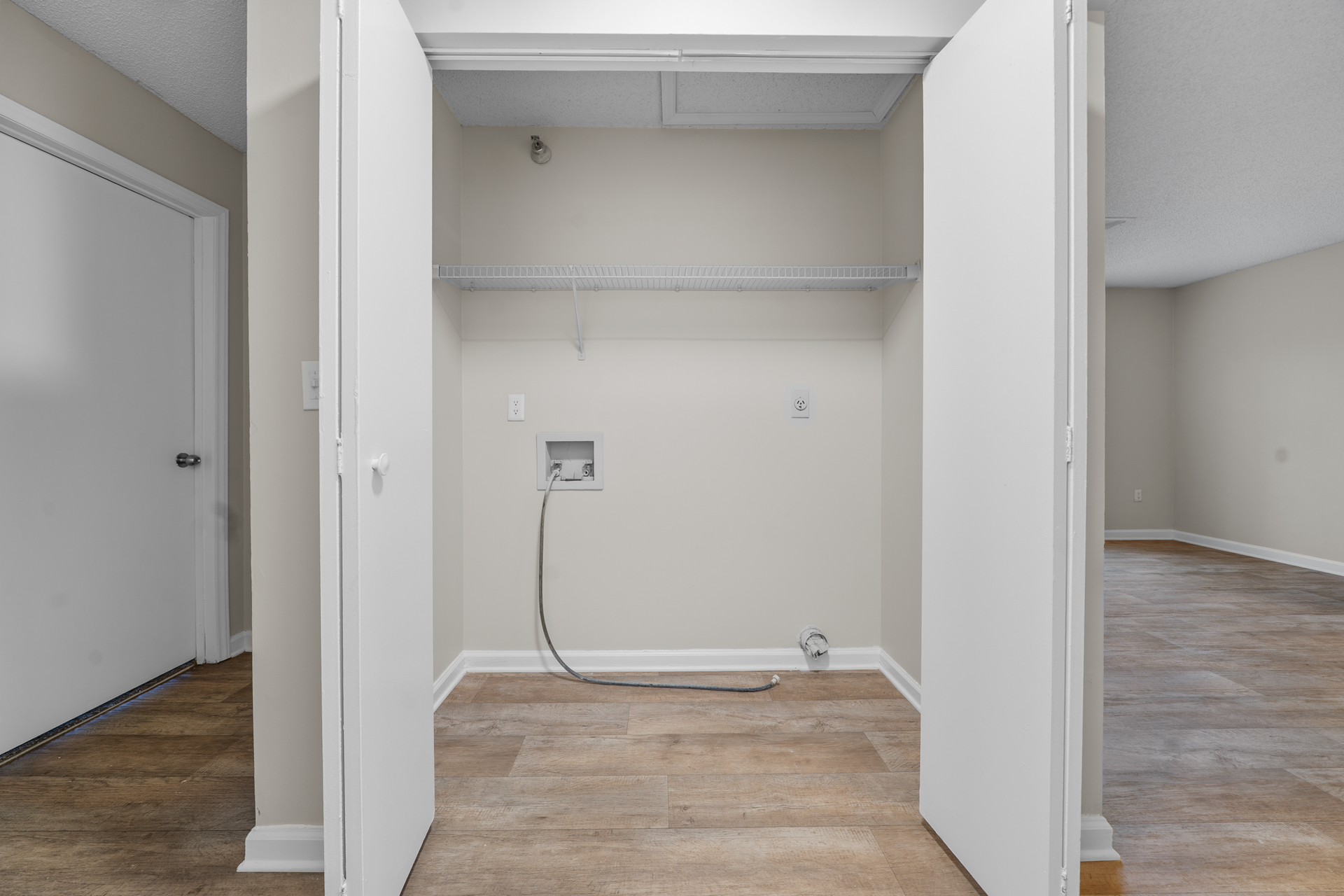 Laundry closet with open white doors, wire shelf, and water hookups, tan floor.