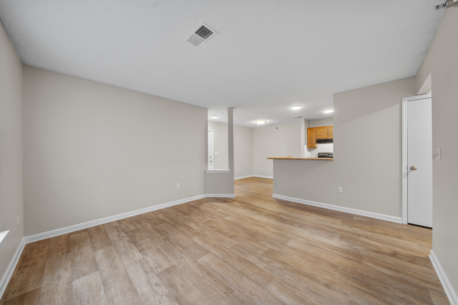 Empty apartment room with wood-look flooring, neutral walls, and a view into the kitchen.