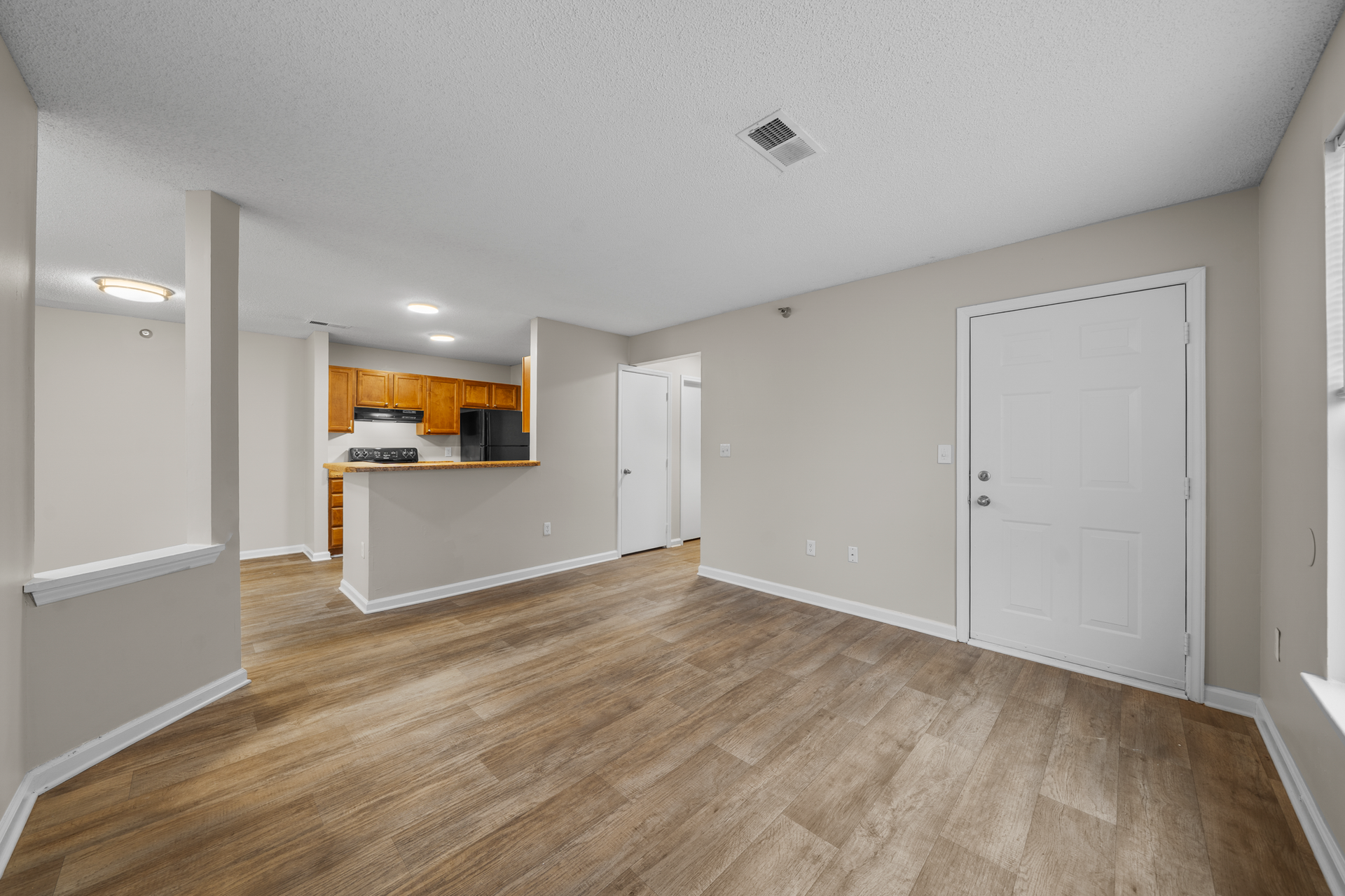 Open-concept living space with wood-look flooring, light walls, and a kitchen visible in the background.