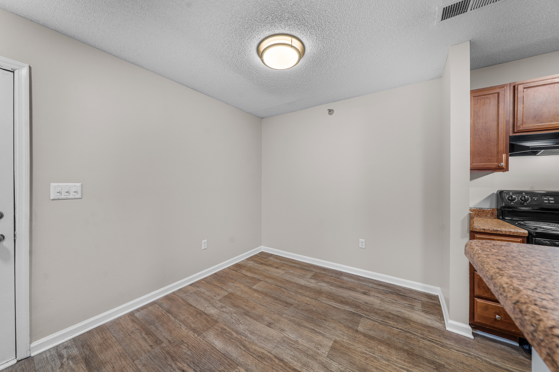 Empty dining area with wood-look flooring, light gray walls, and a recessed ceiling light. Kitchen cabinets on right.