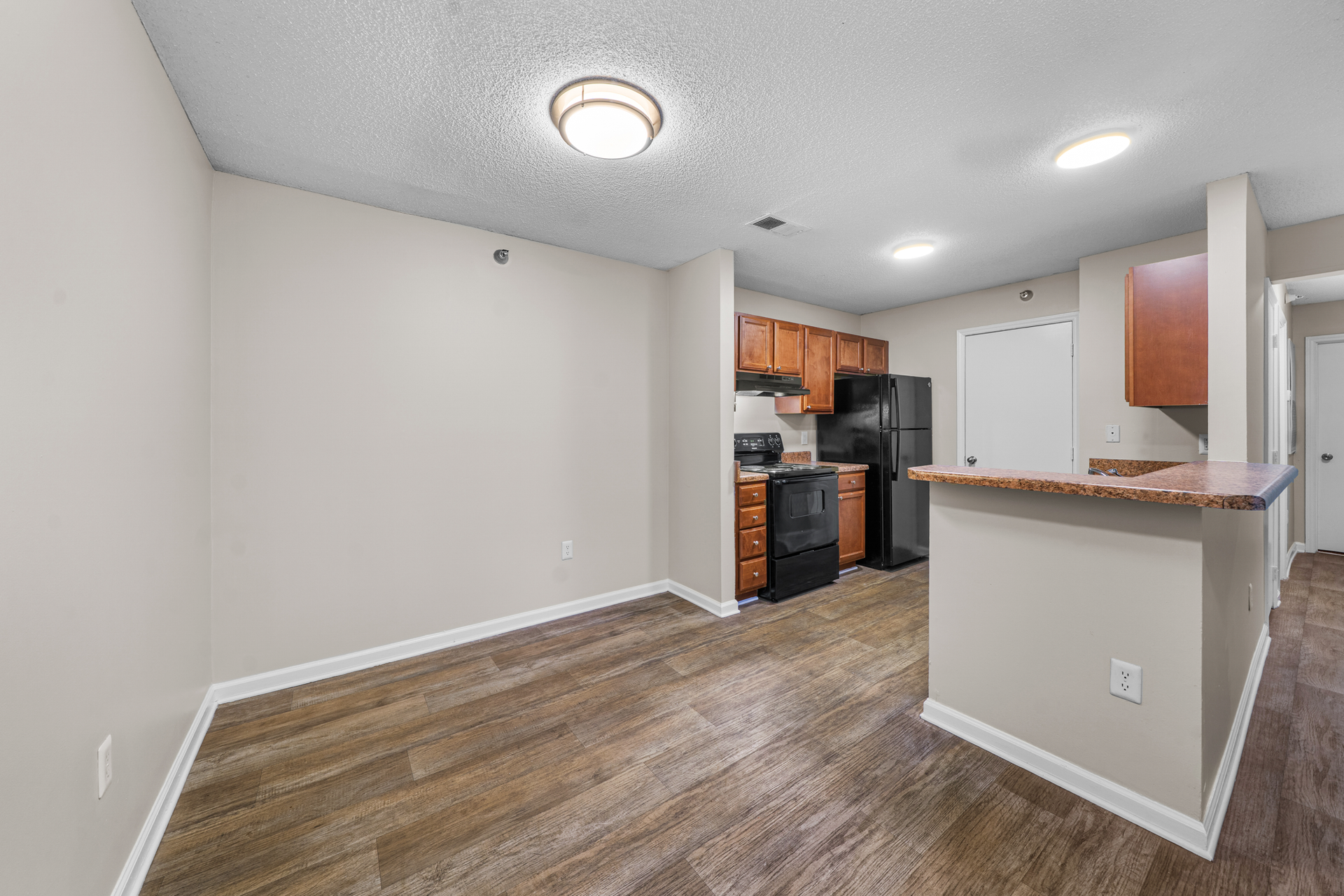 Empty apartment interior with kitchen, wooden floors, beige walls, and brown cabinets.