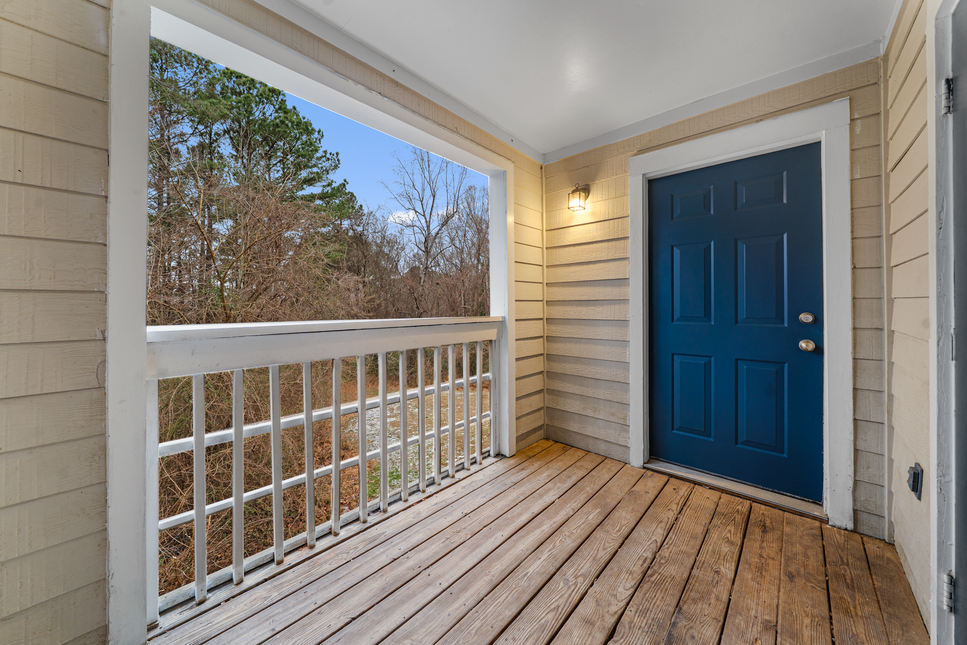 Balcony with wooden floor, white railing, and blue door. View of trees outside.