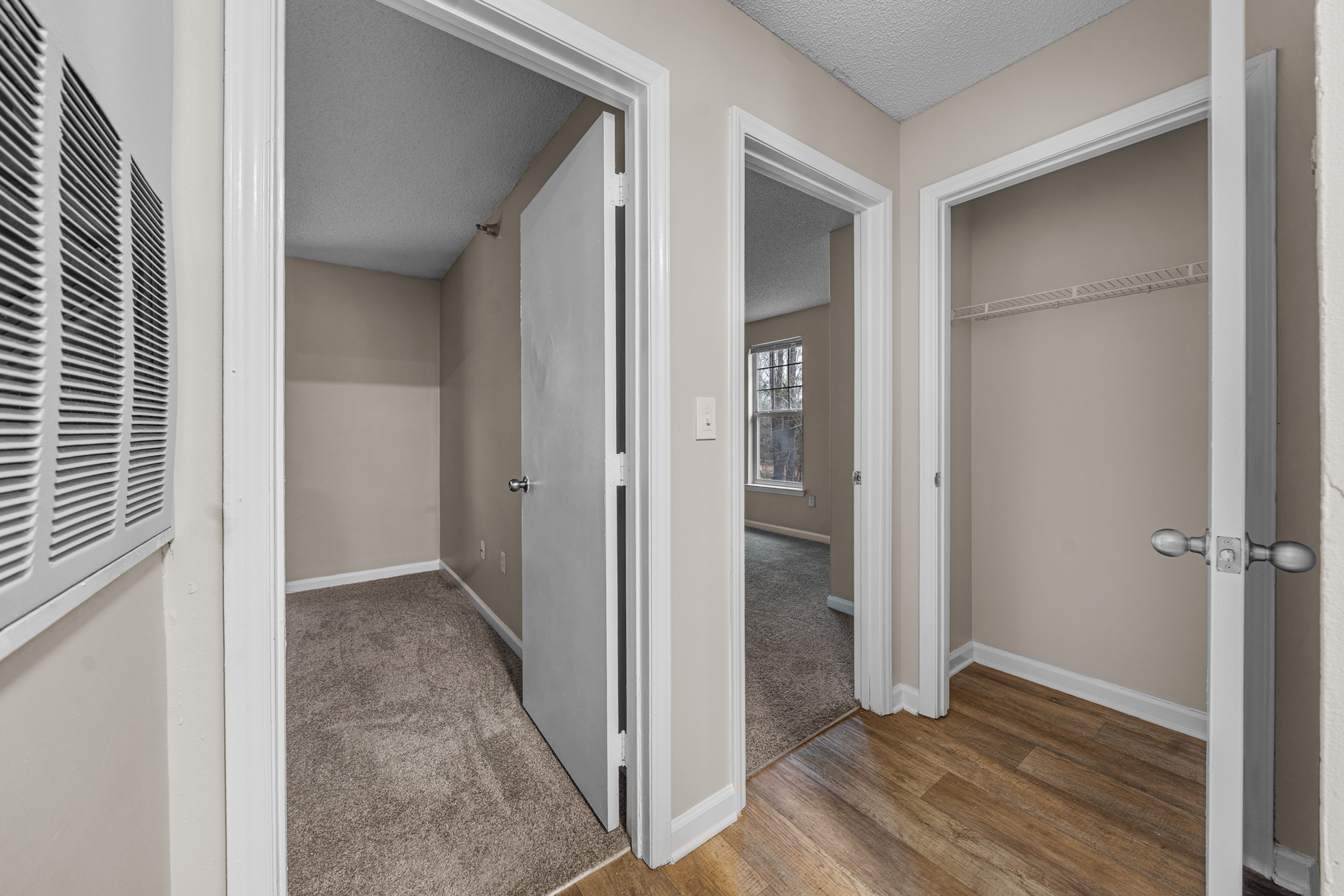 Hallway with three doorways: bedroom, room with window, and closet. Beige walls, wood and carpet floors.