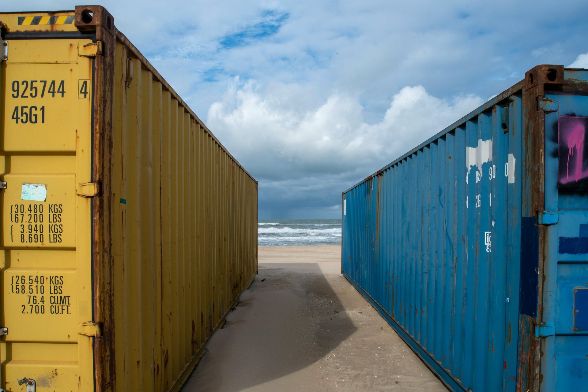 Yellow and blue shipping containers flank a sandy path to the ocean under a cloudy sky.