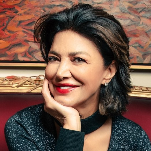 A person with dark hair and bright red lipstick rests their chin on their hand against a red background with art.