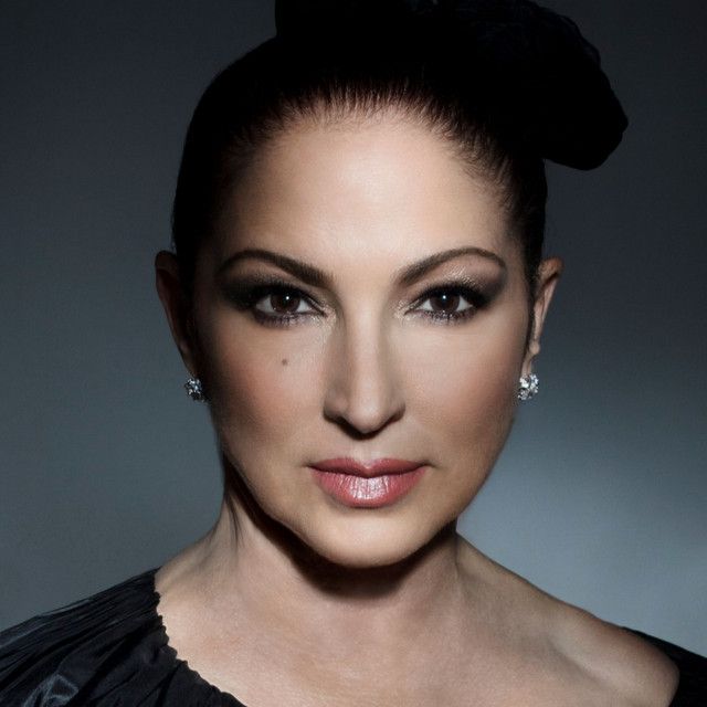 A headshot of a woman with dark hair pulled back, wearing small earrings and a black garment against a dark background.