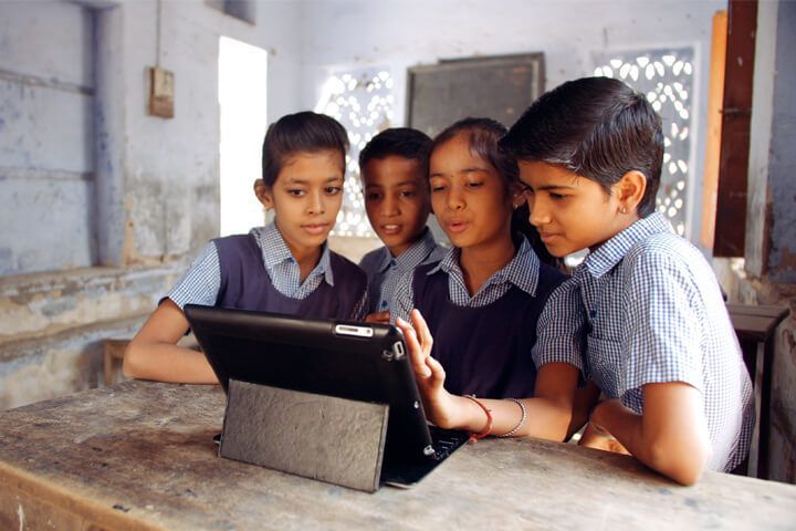 Four students in school uniforms sitting at a wooden desk and looking at a tablet together in a classroom.