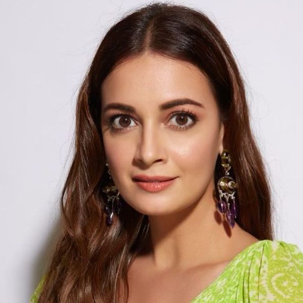 A woman with long brown hair, wearing dangling purple earrings and a green patterned top, looking directly at the camera.