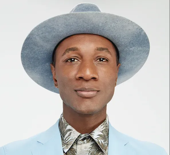 A person wearing a light blue hat and matching suit jacket over a patterned shirt, looking forward against a white background.
