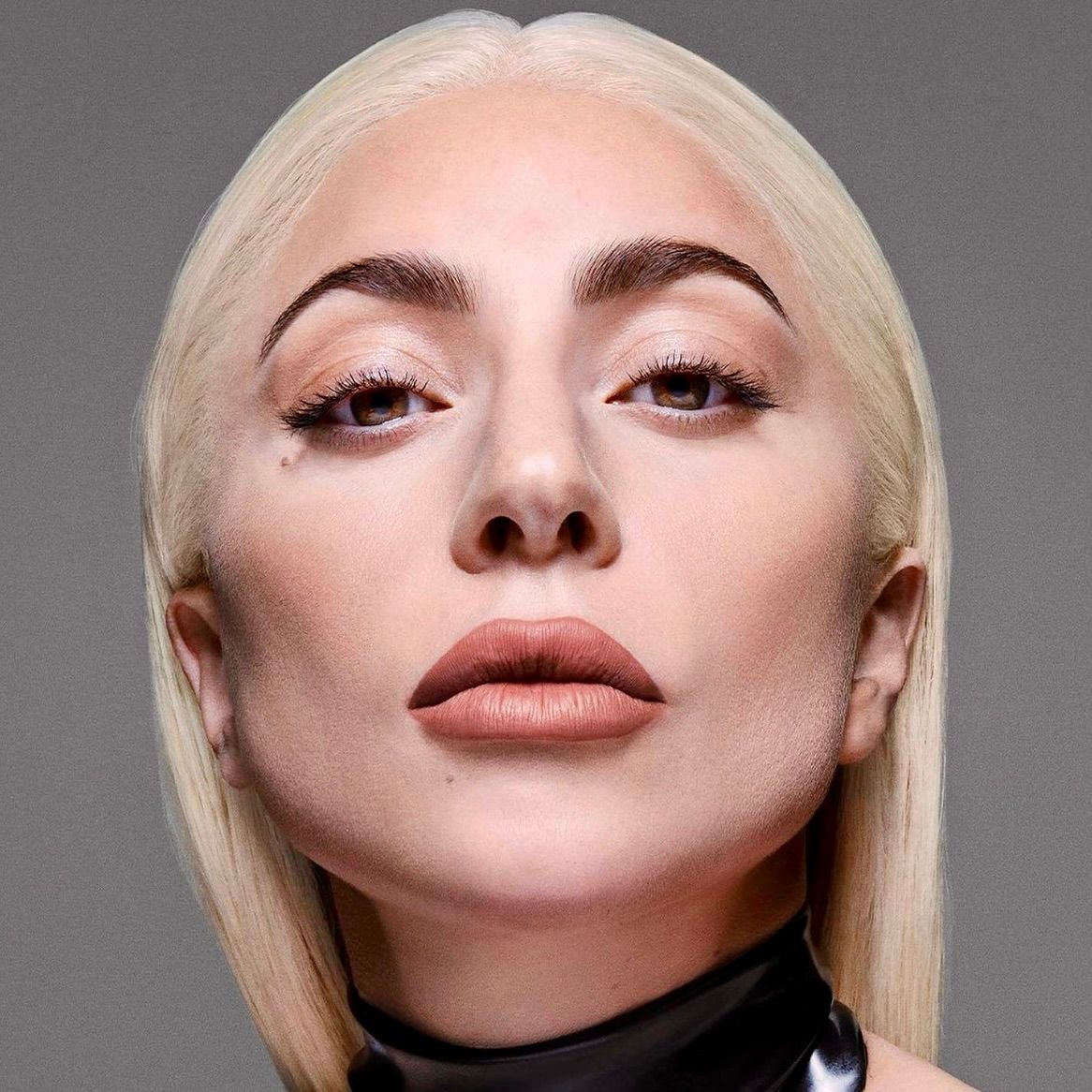 Close-up portrait of Lady Gaga with platinum blonde hair, neutral makeup, and a black collar against a gray background.