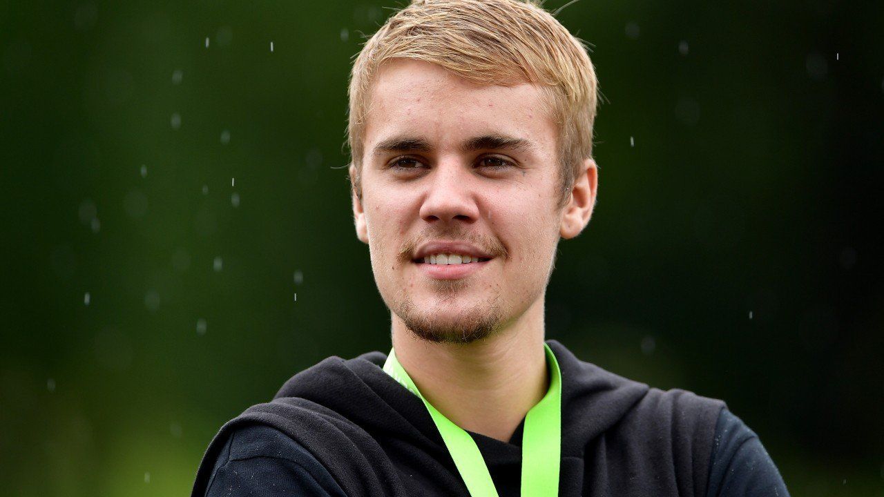 Justin Bieber smiling in a black hoodie against a blurred green background.