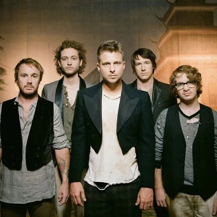 Five men posing for a group portrait against a textured wall, dressed in casual, layered outfits and blazers.