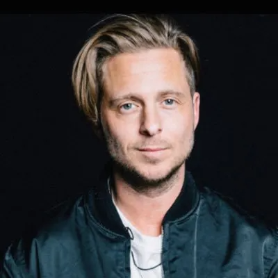 A head-and-shoulders portrait of a person with light brown hair in a dark bomber jacket against a plain black background.