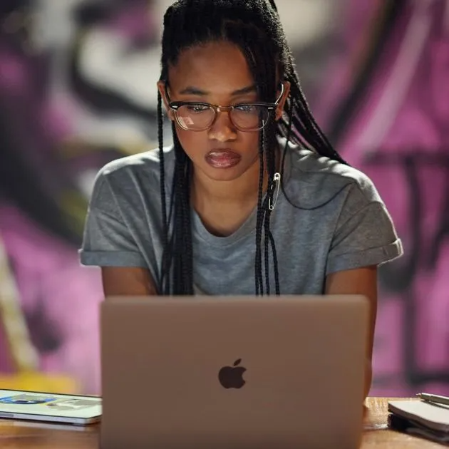 A person with braided hair wearing glasses focuses intently on an open laptop in front of a purple background.