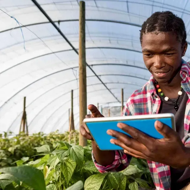 A person in a plaid shirt stands in a greenhouse, smiling while using a tablet to monitor lush green plants.