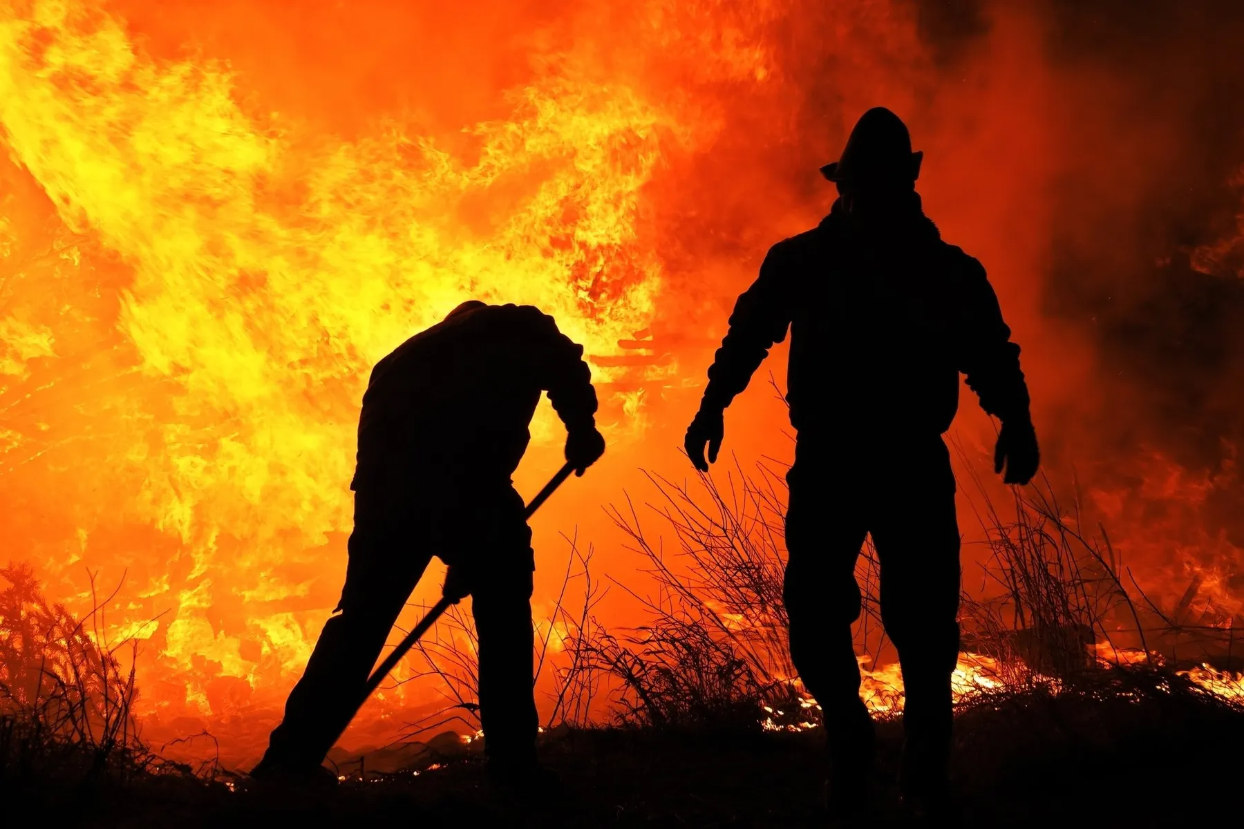 Two firefighters in silhouette working against a large, bright orange wildfire in the background.