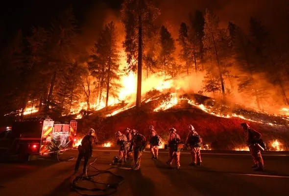 Firefighters in protective gear stand near a fire truck as flames consume a dark, forested hillside at night.