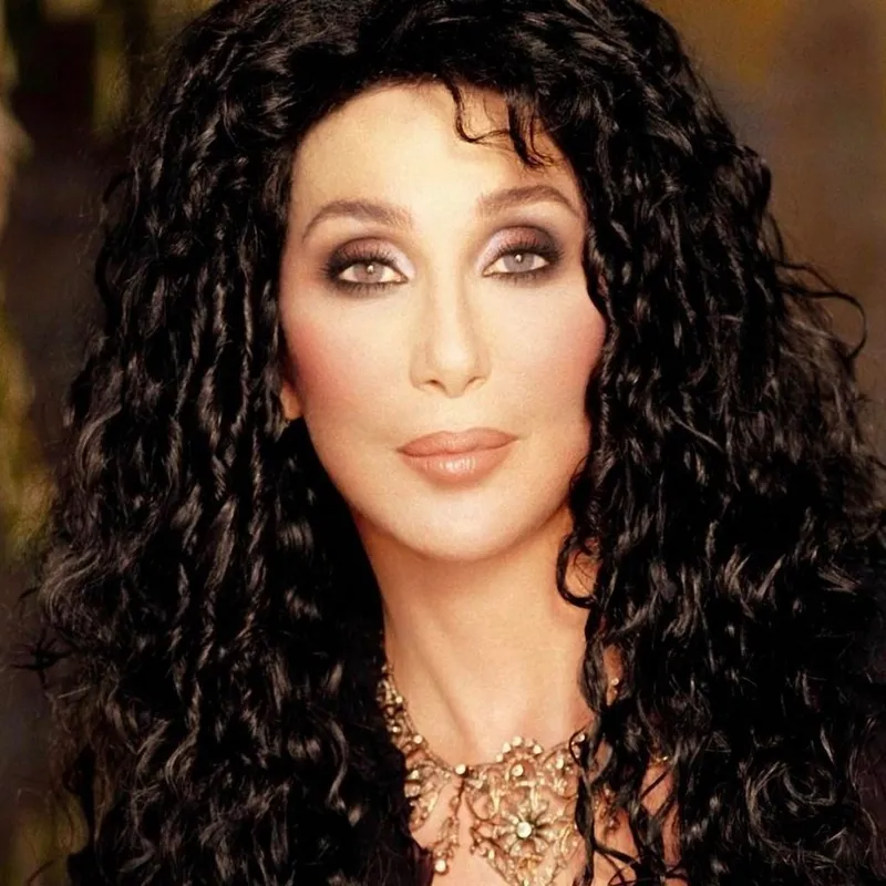 A portrait of Cher with long, curly black hair, wearing smoky eye makeup and a decorative gold-toned necklace.
