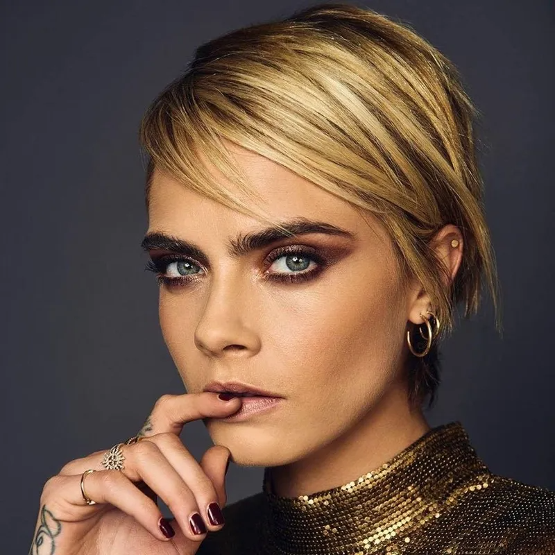 Close-up portrait of Cara Delevingne with blonde hair, dark eye makeup, wearing a gold top, with a hand to her mouth.