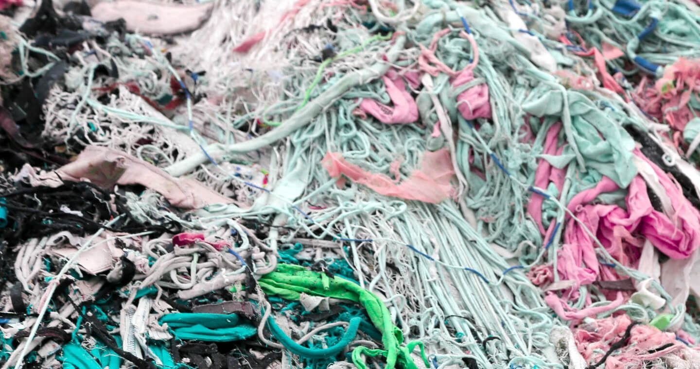 A large pile of shredded, multicolored textile waste featuring strands of pink, light blue, green, and black fabric.