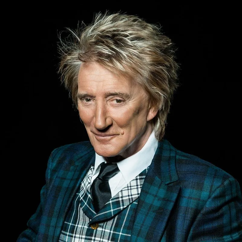 Rod Stewart posing against a black background, wearing a dark green plaid blazer and a black tie over a white shirt.