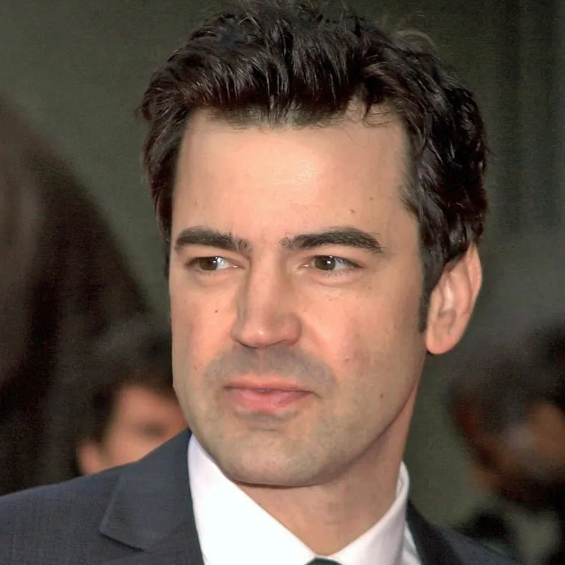 A person with dark hair and a serious expression, wearing a dark suit jacket and white collared shirt.