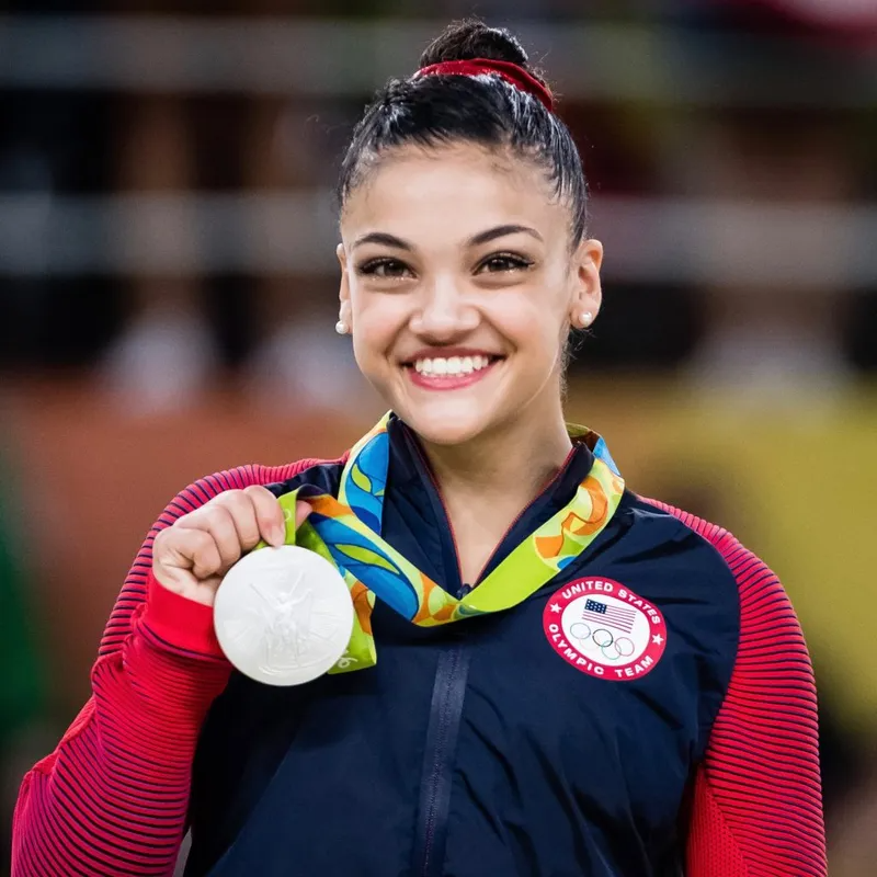A smiling gymnast wearing a United States Olympic team jacket holds up a silver medal.