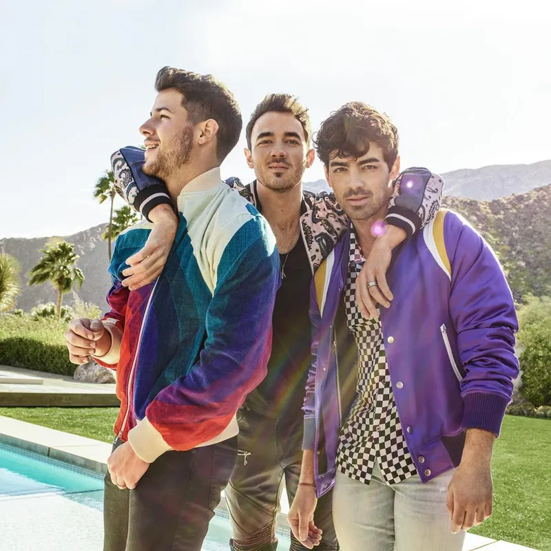 The Jonas Brothers stand together outdoors near a pool, wearing colorful jackets and posing for a bright, sunny portrait.