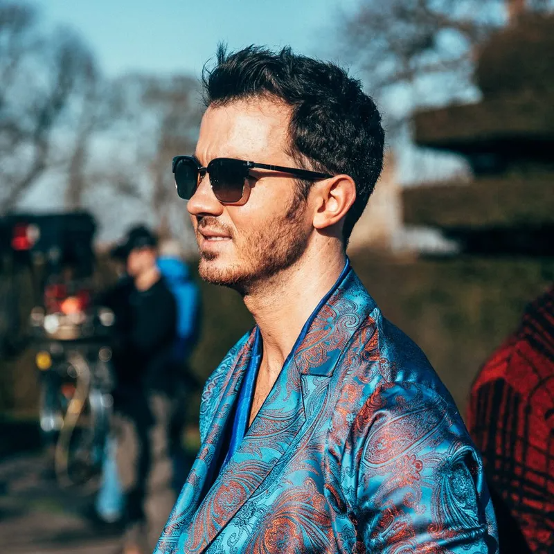 A person in a blue paisley blazer and sunglasses looks to the side outdoors, with a blurred film set in the background.