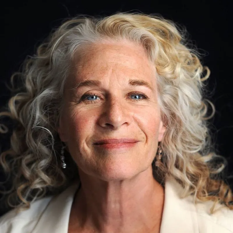 A portrait of a person with curly, shoulder-length blonde hair and blue eyes, smiling gently against a dark background.