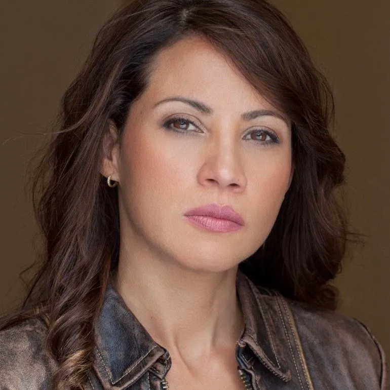 A person with dark hair and a serious expression, wearing a brown leather jacket against a plain background.